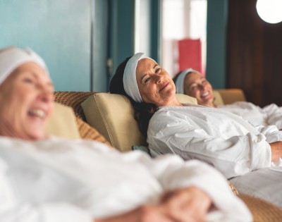 Three women in white robes and headbands relax on spa lounges, smiling contentedly. The setting is tranquil, conveying relaxation and happiness.
