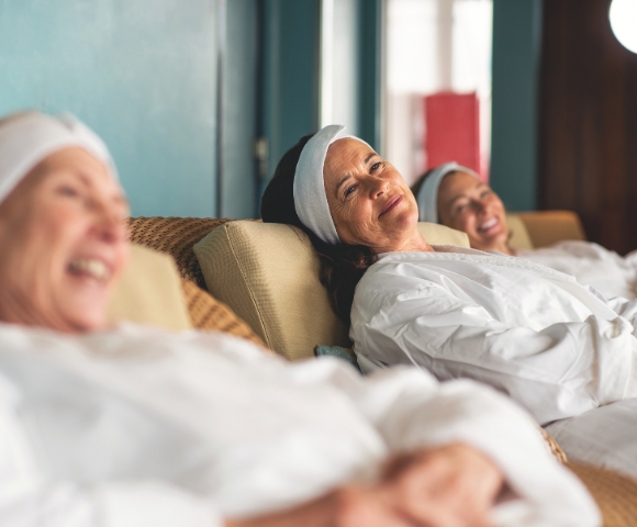 Three women in white robes and headbands relax on spa lounges, smiling contentedly. The setting is tranquil, conveying relaxation and happiness.