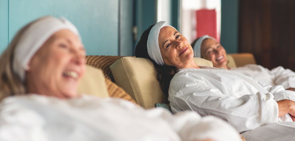 Three women in white robes and headbands relax on spa lounges, smiling contentedly. The setting is tranquil, conveying relaxation and happiness.