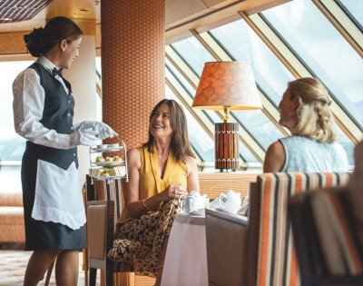 A waitress in a black vest and white gloves serves afternoon tea to two women seated at a table in a sunlit, elegant room. The scene exudes warmth and hospitality.