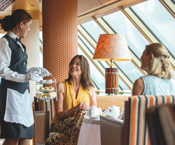 A waitress in a black vest and white gloves serves afternoon tea to two women seated at a table in a sunlit, elegant room. The scene exudes warmth and hospitality.