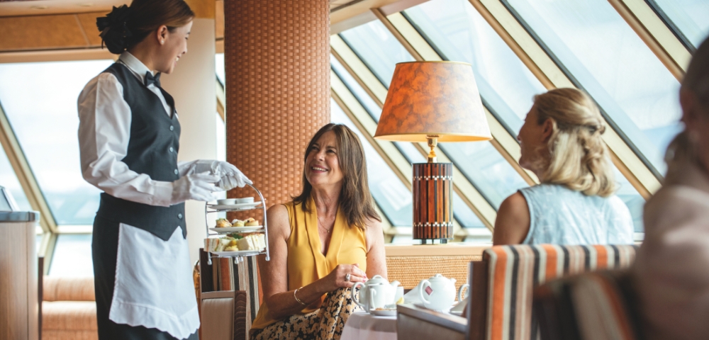 A waitress in a black vest and white gloves serves afternoon tea to two women seated at a table in a sunlit, elegant room. The scene exudes warmth and hospitality.