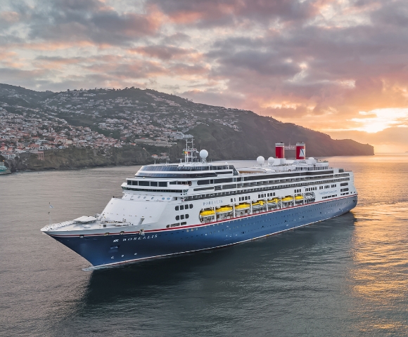 A large blue and white cruise ship sails near an island with mountains at sunset. The sky is filled with colorful clouds, creating a serene and majestic scene.
