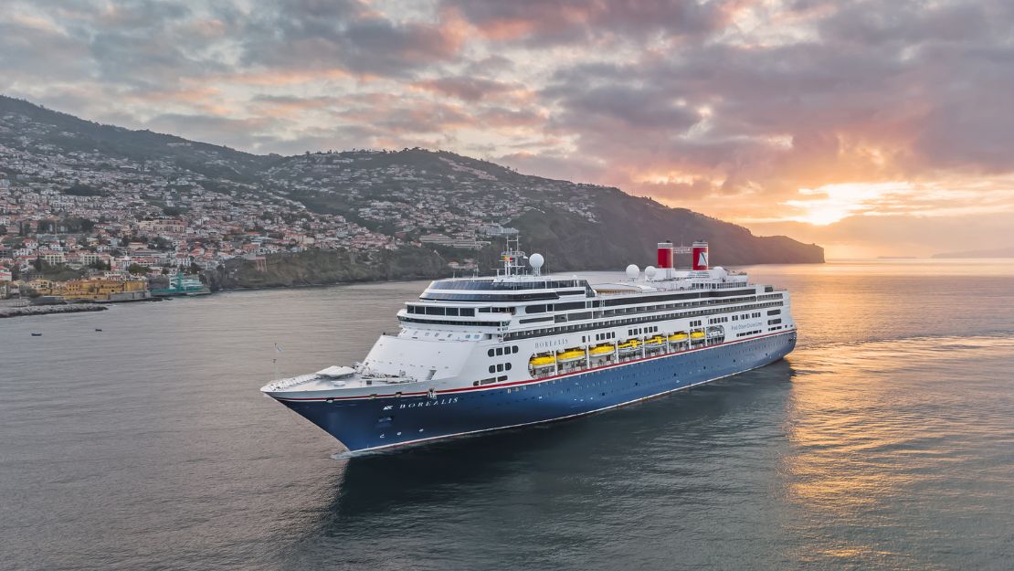 A large blue and white cruise ship sails near an island with mountains at sunset. The sky is filled with colorful clouds, creating a serene and majestic scene.