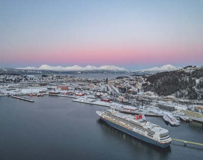 A large cruise ship docked in a snowy port city at dusk with snow-covered hills and mountains in the background under a pastel pink and blue sky.