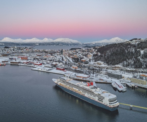 A large cruise ship docked in a snowy port city at dusk with snow-covered hills and mountains in the background under a pastel pink and blue sky.
