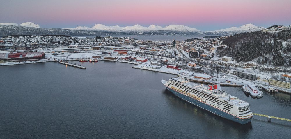 A large cruise ship docked in a snowy port city at dusk with snow-covered hills and mountains in the background under a pastel pink and blue sky.