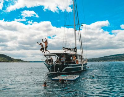 Two people joyfully jump off a sailboat into the ocean, while others swim nearby. The scene is set under a vibrant blue sky with fluffy clouds, evoking fun and adventure.