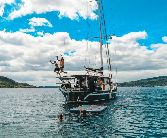 Two people joyfully jump off a sailboat into the ocean, while others swim nearby. The scene is set under a vibrant blue sky with fluffy clouds, evoking fun and adventure.