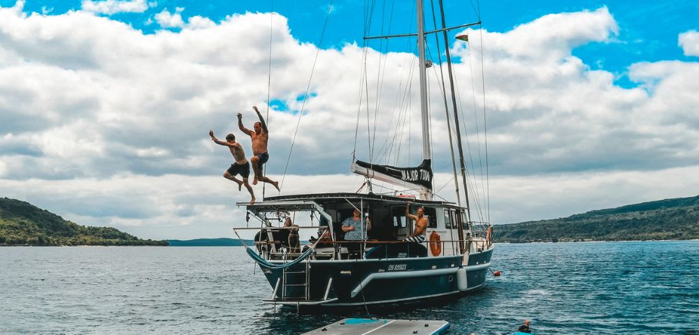 Two people joyfully jump off a sailboat into the ocean, while others swim nearby. The scene is set under a vibrant blue sky with fluffy clouds, evoking fun and adventure.