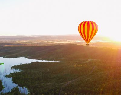 A vibrant red and yellow hot air balloon floats peacefully over a lush, green landscape at sunrise. A serene lake reflects the glowing morning light.