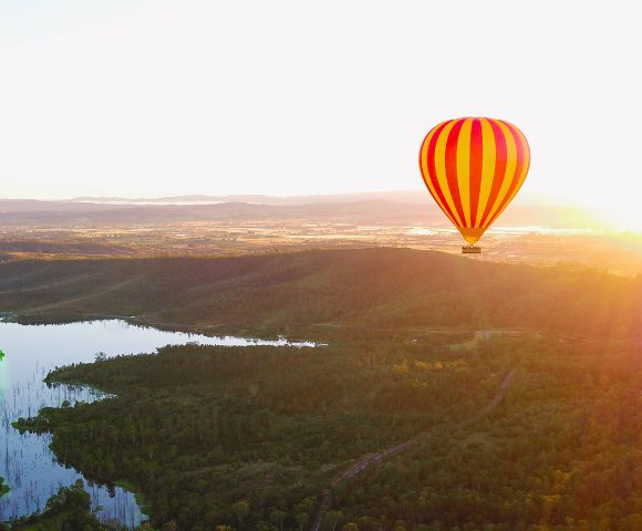 A vibrant red and yellow hot air balloon floats peacefully over a lush, green landscape at sunrise. A serene lake reflects the glowing morning light.