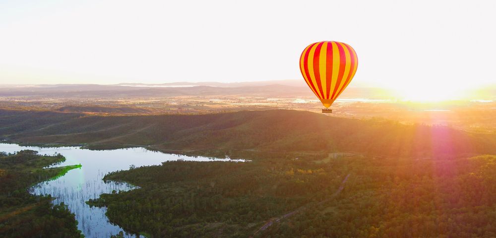 A vibrant red and yellow hot air balloon floats peacefully over a lush, green landscape at sunrise. A serene lake reflects the glowing morning light.