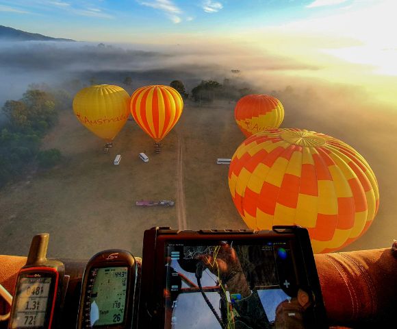 Aerial view from a hot air balloon cockpit showing four colorful balloons with red, yellow, and orange patterns floating over misty fields at sunrise.