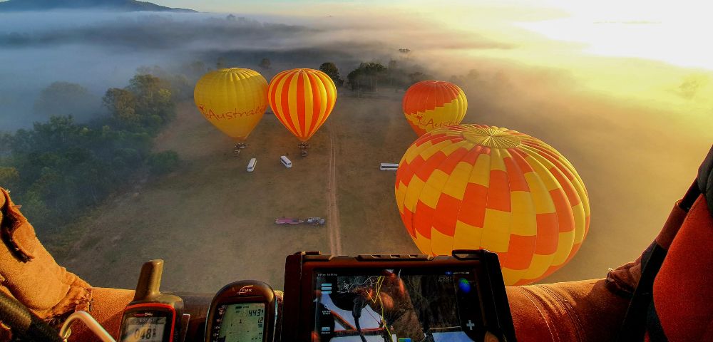 Aerial view from a hot air balloon cockpit showing four colorful balloons with red, yellow, and orange patterns floating over misty fields at sunrise.