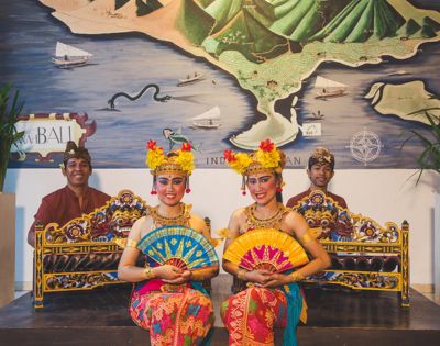 Two Balinese dancers in vibrant traditional attire hold colorful fans, sitting gracefully in front of musicians. A map and instruments are in the background.