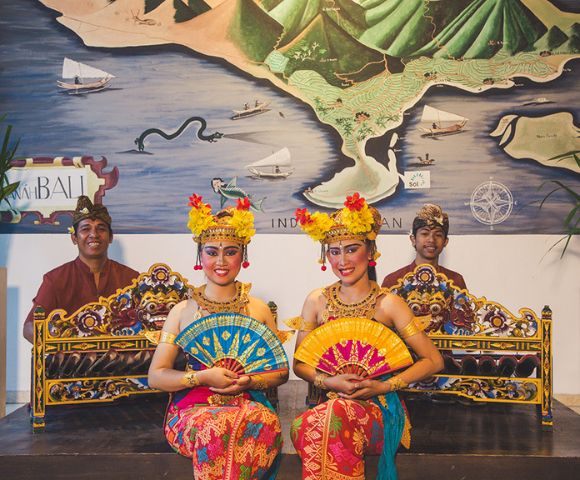 Two Balinese dancers in vibrant traditional attire hold colorful fans, sitting gracefully in front of musicians. A map and instruments are in the background.
