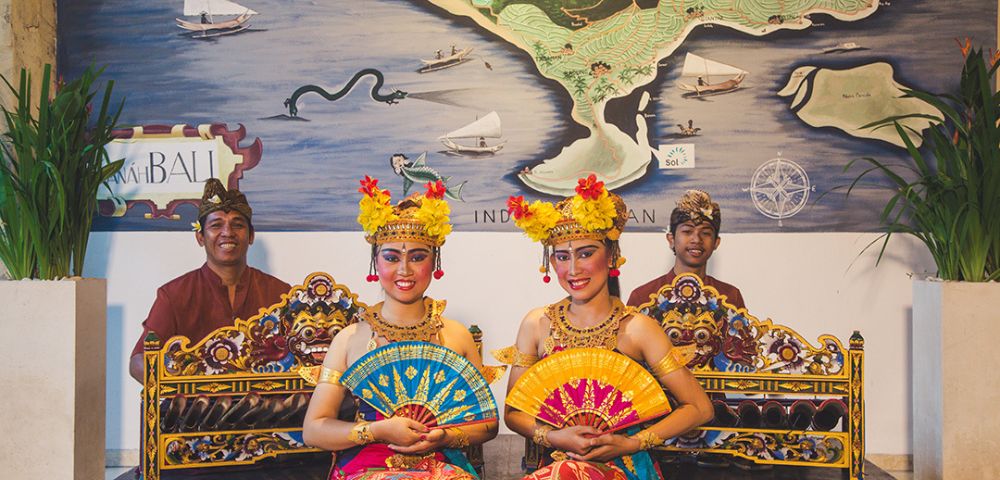 Two Balinese dancers in vibrant traditional attire hold colorful fans, sitting gracefully in front of musicians. A map and instruments are in the background.