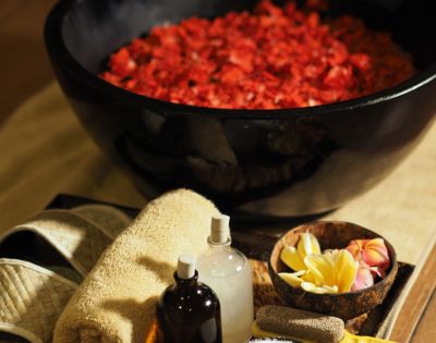 A luxurious spa setting featuring a large black bowl of red rose petals, surrounded by slippers, towels, essential oil bottles, and tropical flowers. Relaxing ambiance.