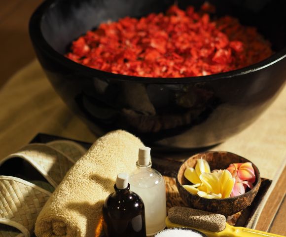 A luxurious spa setting featuring a large black bowl of red rose petals, surrounded by slippers, towels, essential oil bottles, and tropical flowers. Relaxing ambiance.