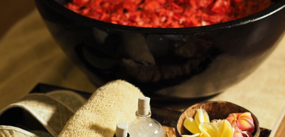 A luxurious spa setting featuring a large black bowl of red rose petals, surrounded by slippers, towels, essential oil bottles, and tropical flowers. Relaxing ambiance.