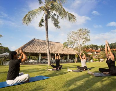 Four people practice yoga under a tall palm tree on a sunny day. They sit cross-legged on mats in front of a thatched-roof hut, arms raised peacefully.