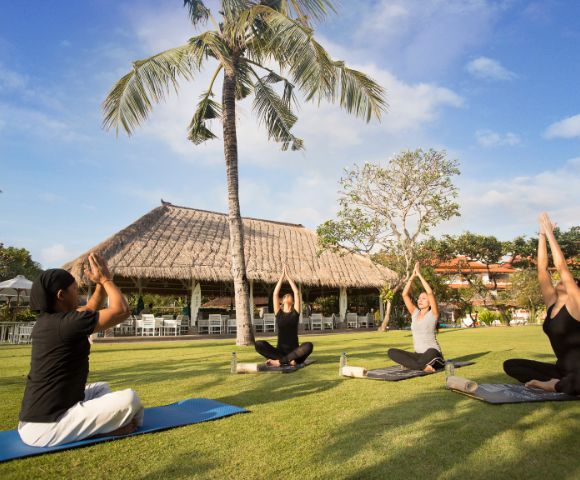 Four people practice yoga under a tall palm tree on a sunny day. They sit cross-legged on mats in front of a thatched-roof hut, arms raised peacefully.