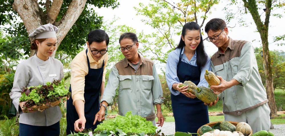 A diverse group of five people stands together outdoors, smiling and examining fresh vegetables and greens in baskets, conveying a sense of community and harvest.