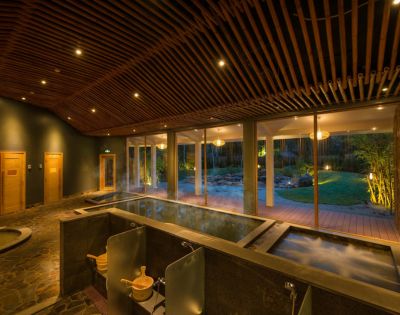 Cozy spa interior with warm lighting, featuring two steaming hot tubs, wooden ceiling, large windows, and a garden view, exuding relaxation and tranquility.