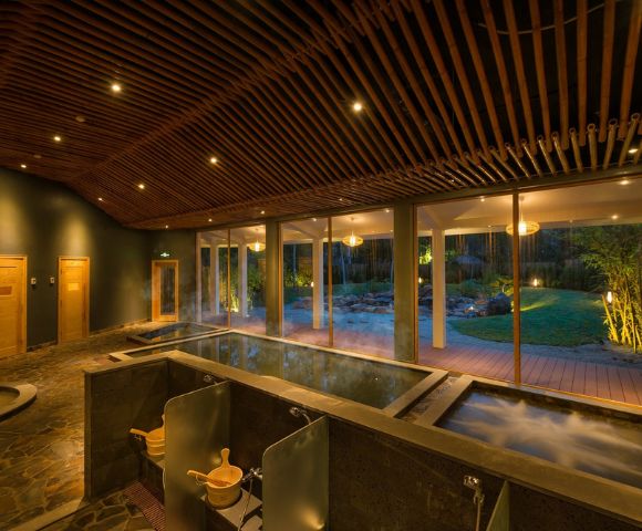Cozy spa interior with warm lighting, featuring two steaming hot tubs, wooden ceiling, large windows, and a garden view, exuding relaxation and tranquility.