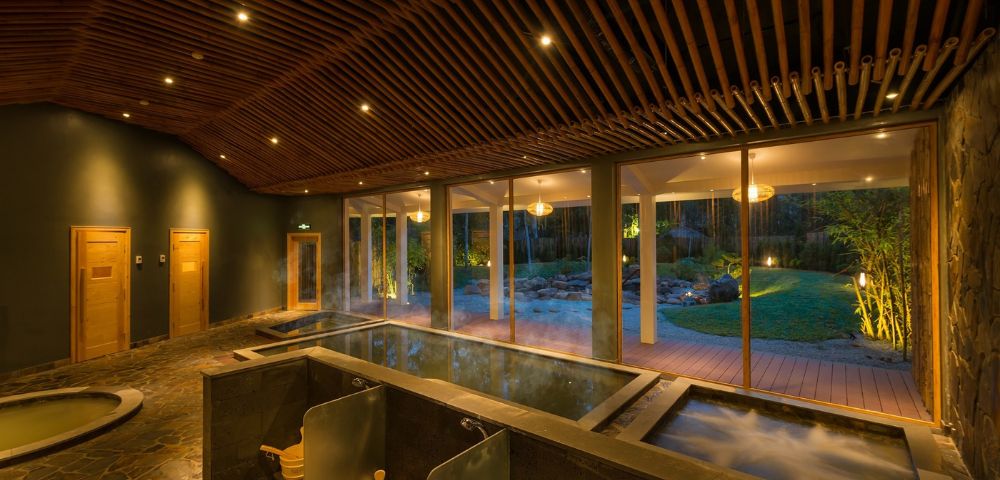 Cozy spa interior with warm lighting, featuring two steaming hot tubs, wooden ceiling, large windows, and a garden view, exuding relaxation and tranquility.