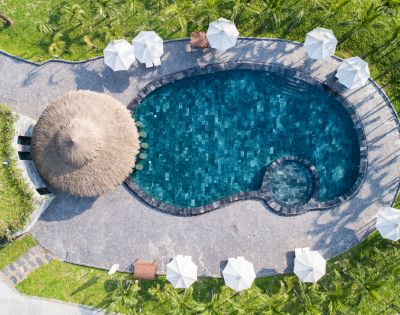 Aerial view of an oval pool with deep blue water, surrounded by white umbrellas and palm trees. A thatched-roof hut adds a tropical vibe.