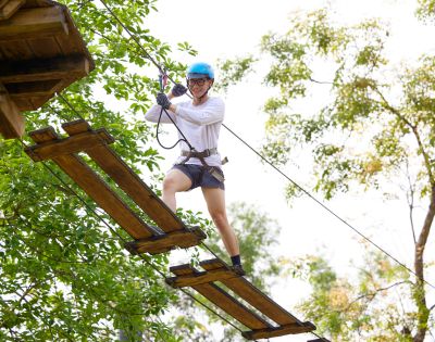 Person in blue helmet and harness crossing a wooden rope bridge high above ground amidst green trees, conveying adventure and excitement.