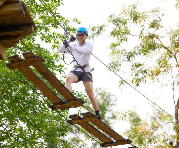 Person in blue helmet and harness crossing a wooden rope bridge high above ground amidst green trees, conveying adventure and excitement.