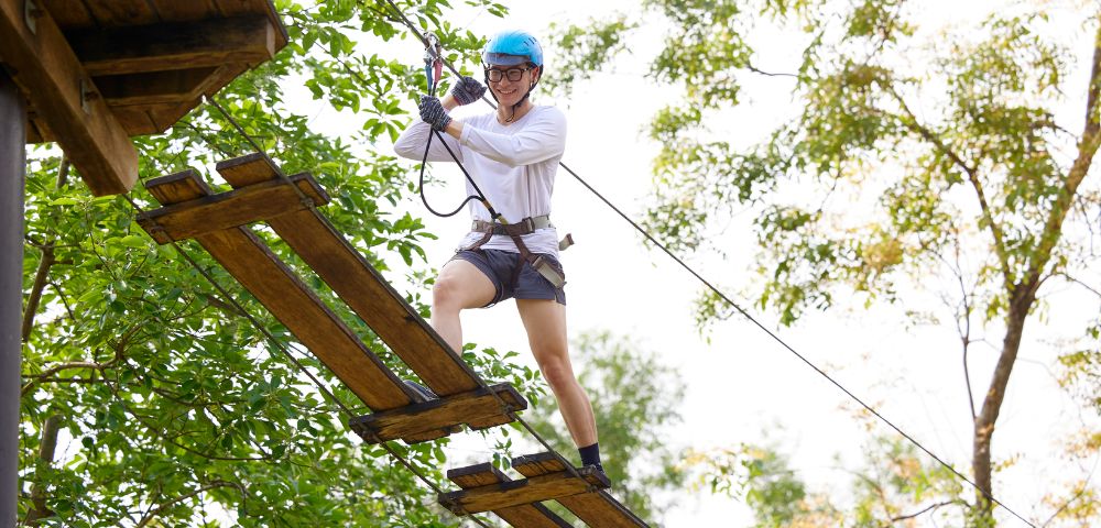 Person in blue helmet and harness crossing a wooden rope bridge high above ground amidst green trees, conveying adventure and excitement.