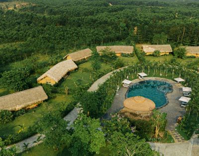 Aerial view of a serene resort with thatched-roof bungalows, a circular pool surrounded by palm trees, and lush greenery, conveying a peaceful, tropical atmosphere.