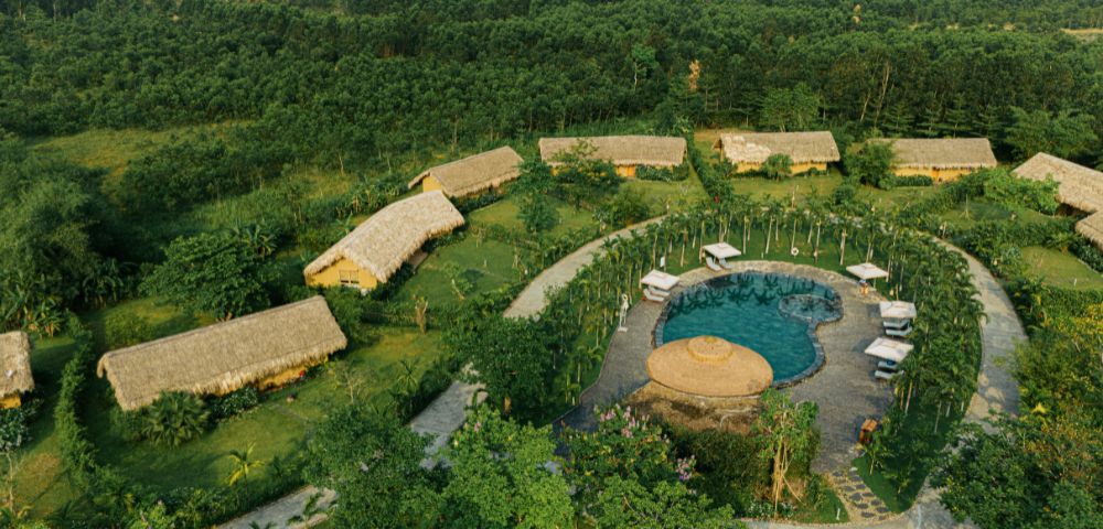 Aerial view of a serene resort with thatched-roof bungalows, a circular pool surrounded by palm trees, and lush greenery, conveying a peaceful, tropical atmosphere.