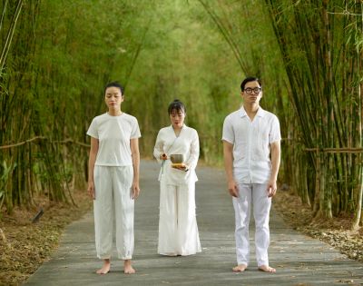 Three individuals in white stand on a serene bamboo path. Two face forward with calm expressions, while one behind holds a bowl, suggesting ritual or meditation.