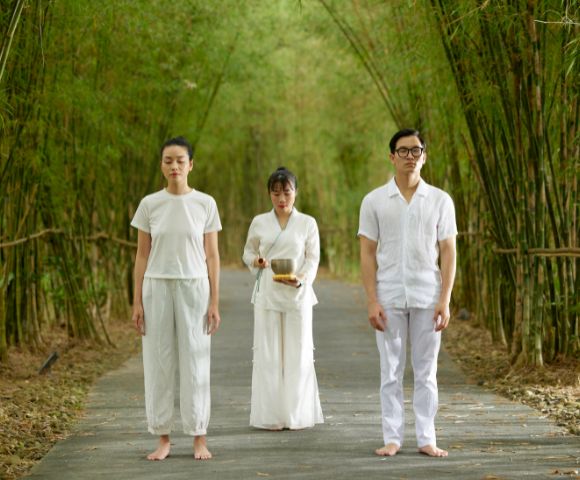 Three individuals in white stand on a serene bamboo path. Two face forward with calm expressions, while one behind holds a bowl, suggesting ritual or meditation.