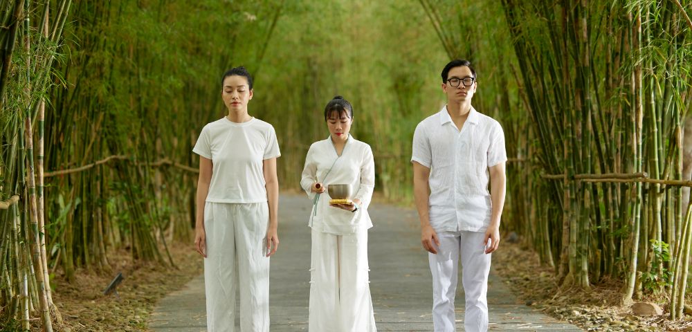Three individuals in white stand on a serene bamboo path. Two face forward with calm expressions, while one behind holds a bowl, suggesting ritual or meditation.