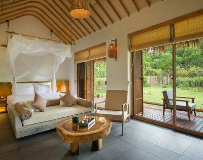 Elegant hotel room with a canopy bed, neutral palette, and wood accents. Large windows open to a lush garden, creating a serene, inviting atmosphere.