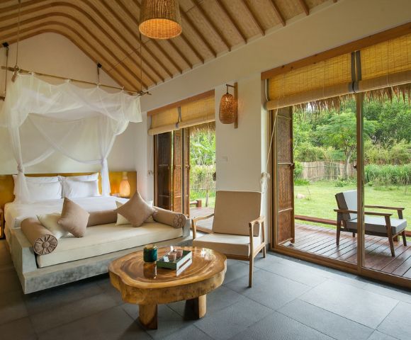 Elegant hotel room with a canopy bed, neutral palette, and wood accents. Large windows open to a lush garden, creating a serene, inviting atmosphere.