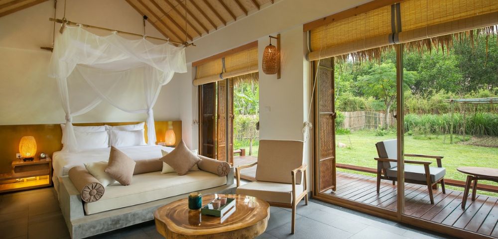 Elegant hotel room with a canopy bed, neutral palette, and wood accents. Large windows open to a lush garden, creating a serene, inviting atmosphere.