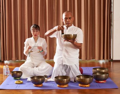 A man and woman in white clothing perform a sound healing session with Tibetan singing bowls on a yoga mat, evoking calm and meditation.