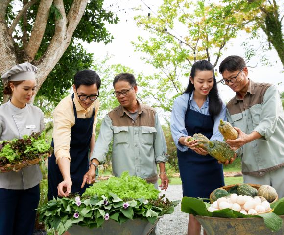 A diverse group of five people stands together outdoors, smiling and examining fresh vegetables and greens in baskets, conveying a sense of community and harvest.