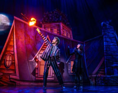 A dramatic stage scene depicts two performers on a rooftop. One raises an arm, conjuring flames. The vibrant lighting enhances a mystical, theatrical atmosphere.