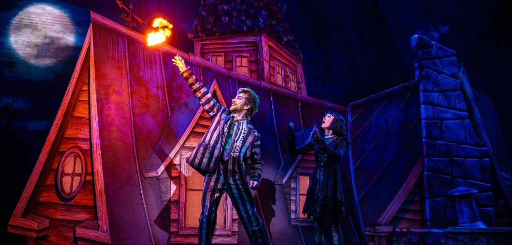 A dramatic stage scene depicts two performers on a rooftop. One raises an arm, conjuring flames. The vibrant lighting enhances a mystical, theatrical atmosphere.