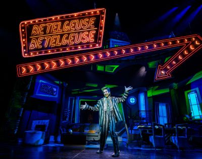 A performer in a striped suit stands on stage with arms outstretched beneath a neon 