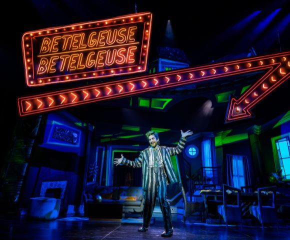 A performer in a striped suit stands on stage with arms outstretched beneath a neon 