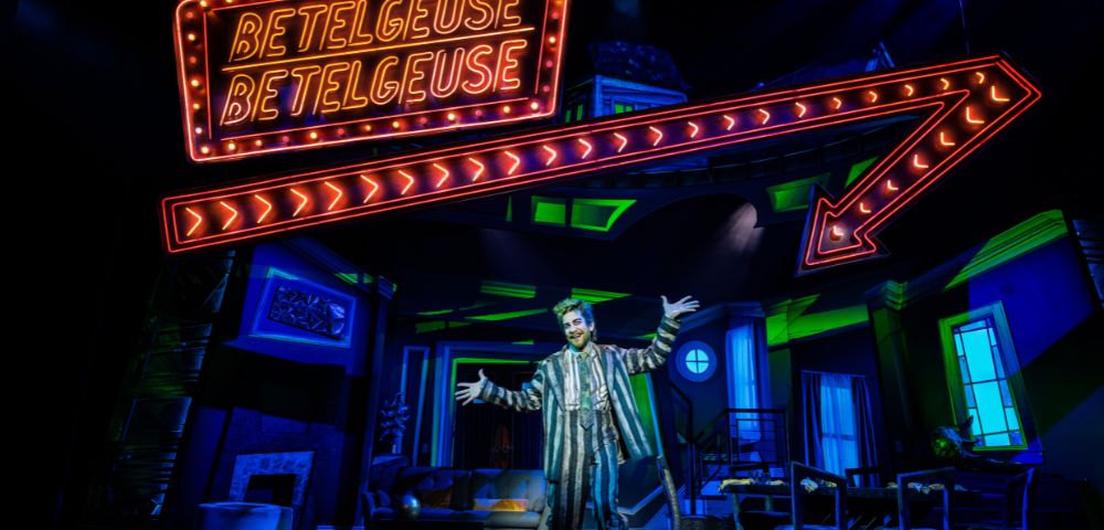 A performer in a striped suit stands on stage with arms outstretched beneath a neon 